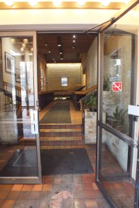 L'intérieur d'une entrée d'un immeuble avec deux portes en vitres. L'une des portes est ouverte. Au-delà des portes se trouvent trois marches qui mènent à un couloir.