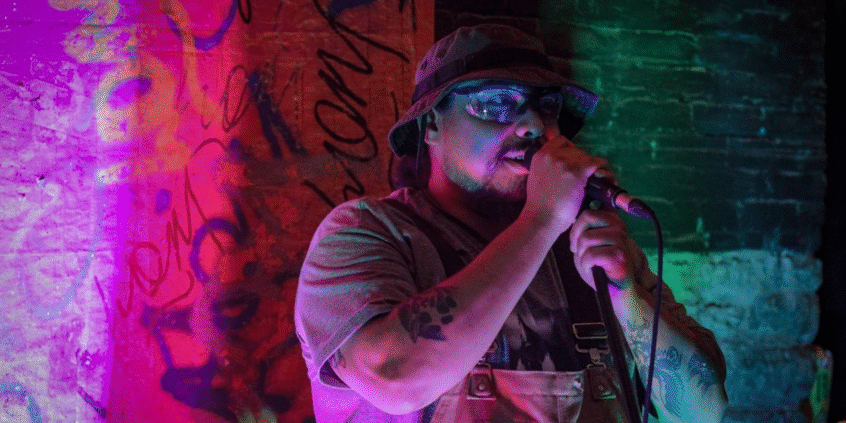 A photo of interdisciplinary artist Pablo Javier Castillo Huerta. They are standing in an alleyway at night, illuminated by neon lights, and holding a microphone to their mouth.