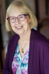 A photo of Judy Murphy, smiling at the camera. She is wearing glasses, a patterned shirt, and a purple cardigan.